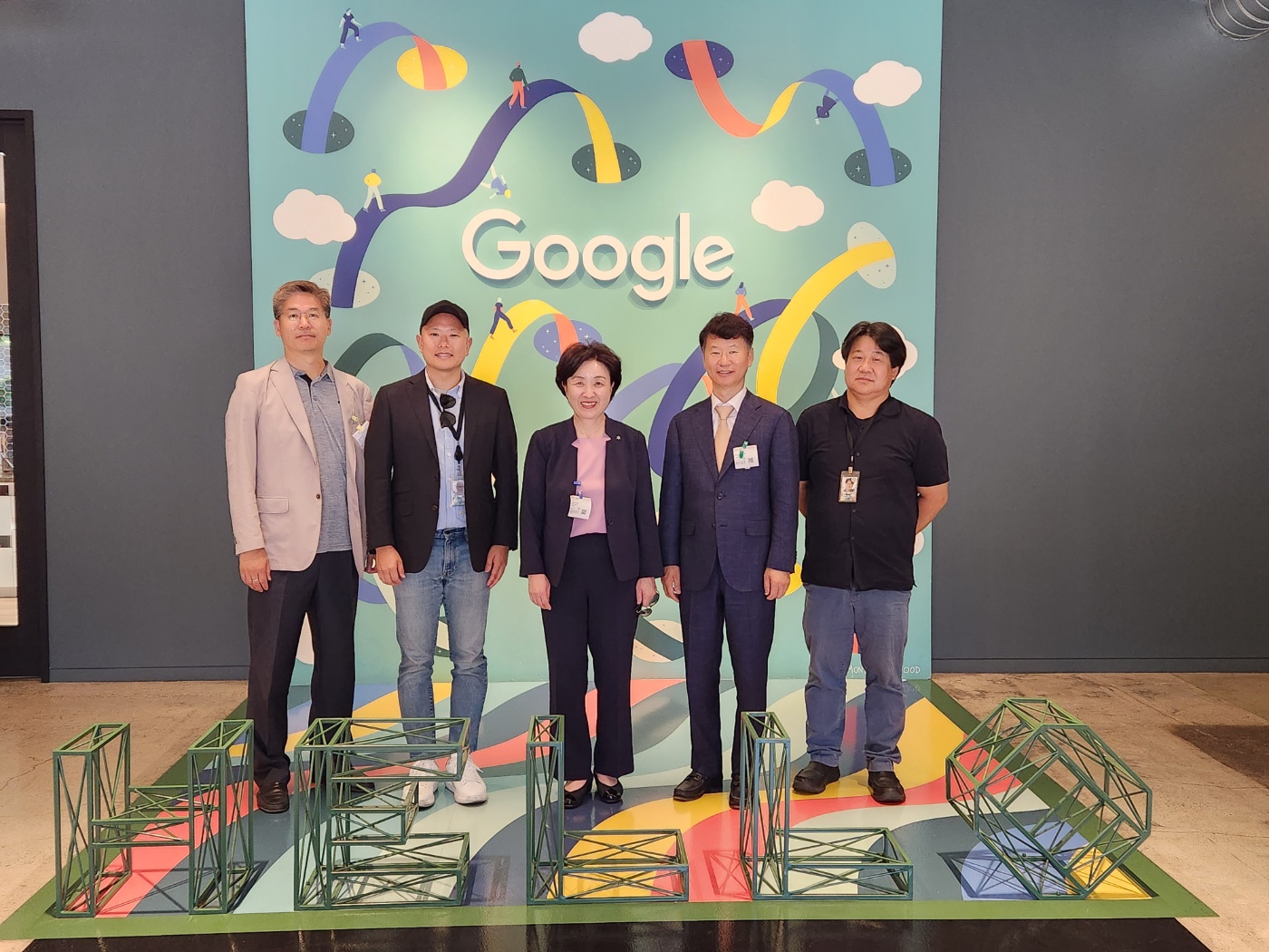 가운데 중년으로 보이는 여성이 서 있고, 좌우로 두 명씩 남성이 서 있다. 뒤로는 Google이라고 써진 글자가 보인다.